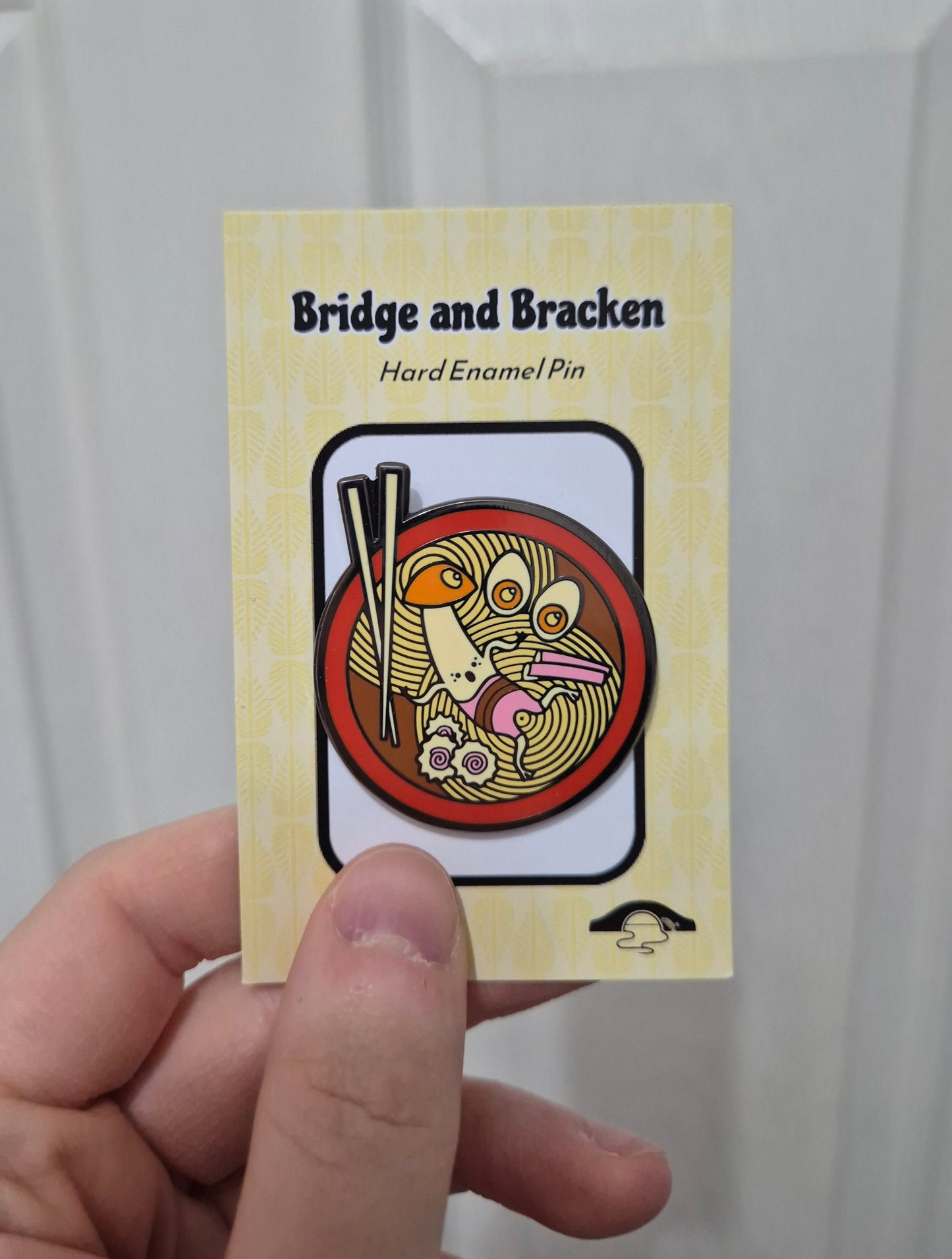 Hand holding a hard enamel pin design featuring a mushroom character, noodles and chopsticks.