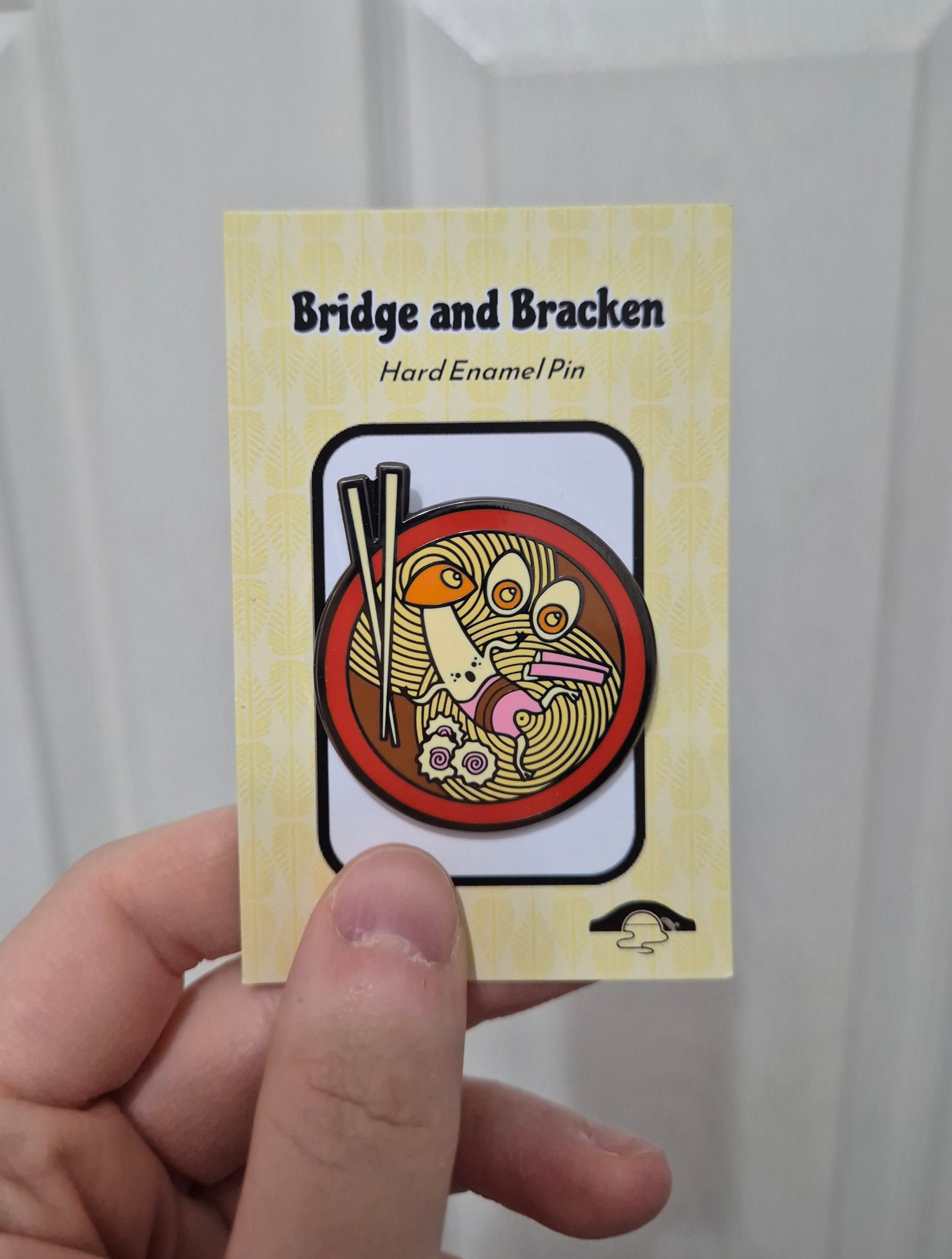 Hand holding a hard enamel pin design featuring a mushroom character, noodles and chopsticks.