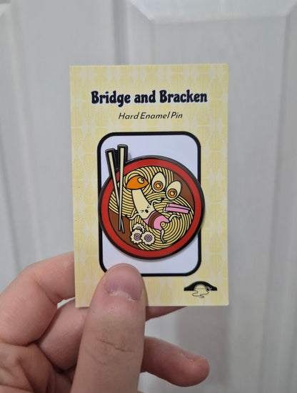 Hand holding a hard enamel pin design featuring a mushroom character, noodles and chopsticks.